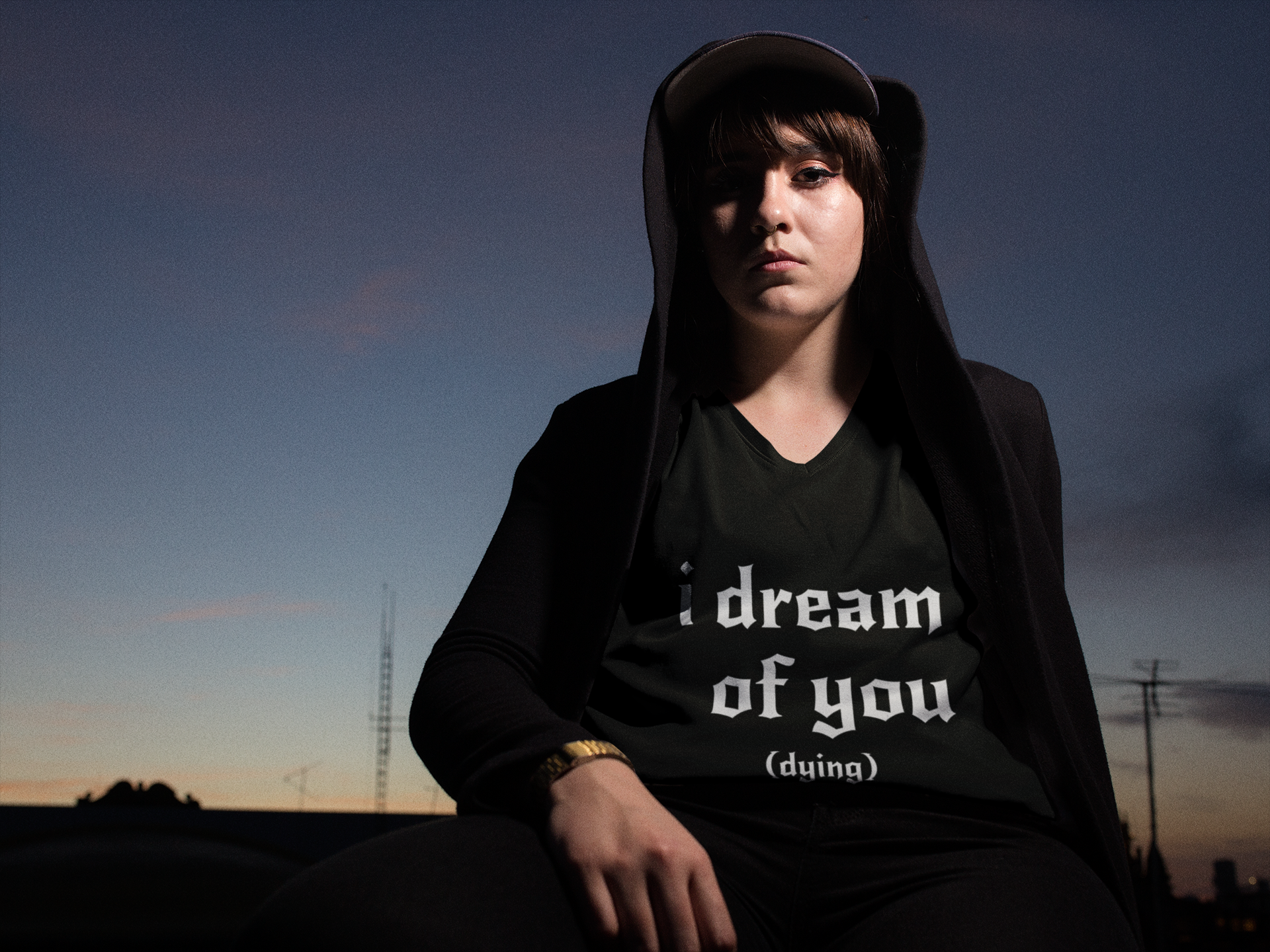 Person wearing a black hoodie with text sitting outdoors at dusk. Text reads I dream of you...dying