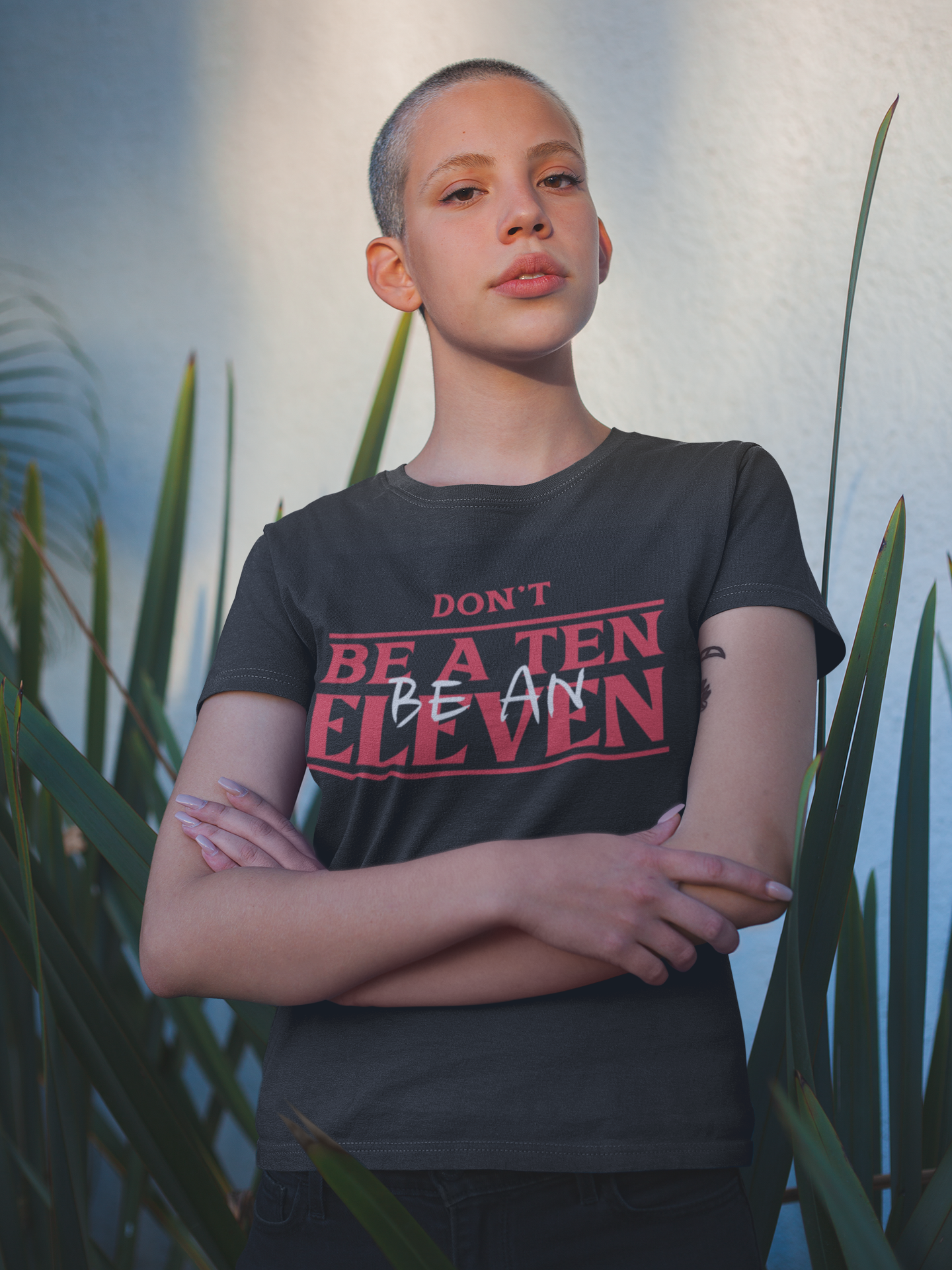 Person wearing a black t-shirt with red text standing in front of plants. Shirt reads Don't be a ten, Be an Eleven. Mock Stranger Things Font